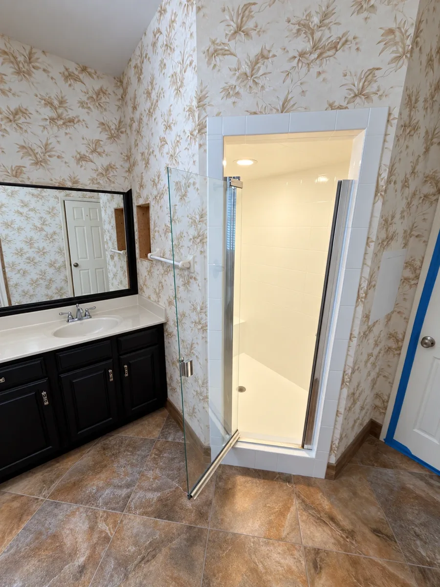 Before: small fiberglass shower stall with floral wallpaper and black vanity in a St. Johns master bathroom