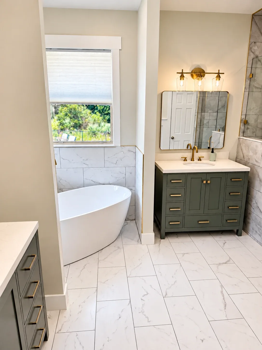 Freestanding soaking tub with marble-look porcelain wainscot and gold trim next to a green vanity, master bathroom remodel in St. Johns FL